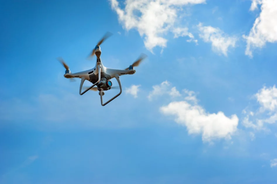 Biały dron z podwieszoną kamerą w trakcie lotu na tle błękitnego nieba