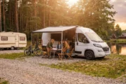 Kamper z rozłożoną markizą i przyczepa kempingowa na leśnym kempingu nad jeziorem, rodzina przygotowuje posiłek - ubezpieczenie kampera i przyczepy kempingowej